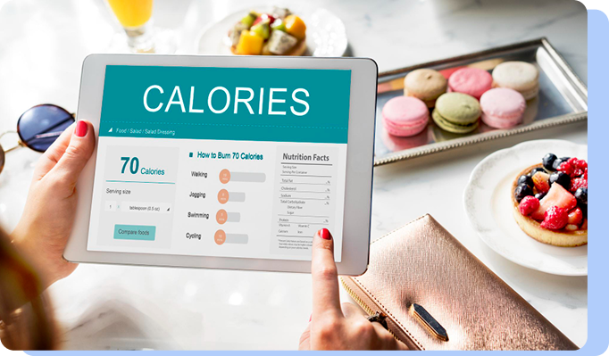 A person holding a digital tablet that displays a "CALORIES" tracking interface with various charts. In the background, a tray of colorful macarons and a fruit tart are visible on a table.