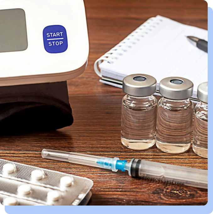 A variety of medical supplies on a wooden table, including three glass vials, a syringe, a blister pack of pills, a notebook, and a digital blood pressure monitor.