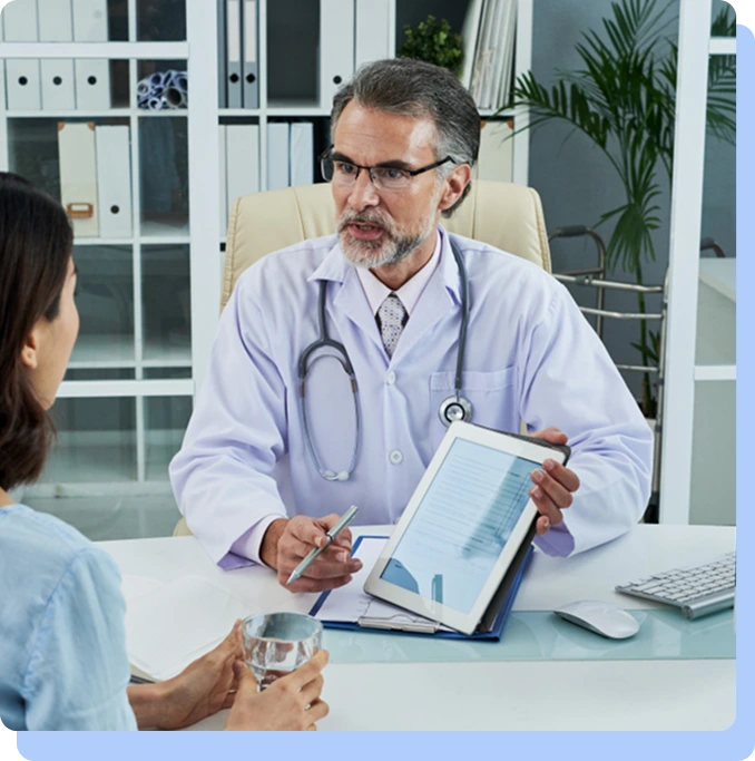 A male doctor in a lab coat sitting at a desk and showing a digital tablet to a patient during a consultation.
