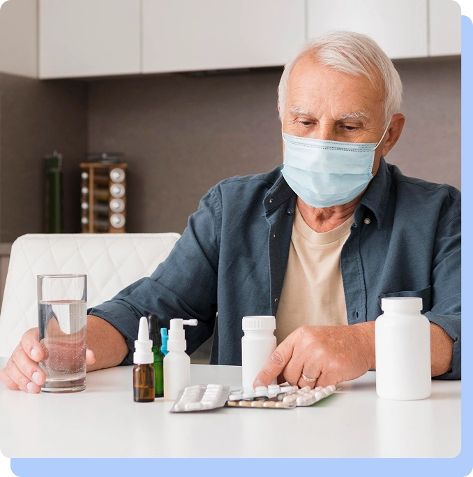 An older man in a face mask looking at various medications and a glass of water on a table.