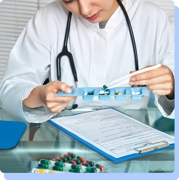 A female healthcare provider in a white lab coat sitting at a desk, carefully organizing small green and blue tablets into a weekly pill organizer.