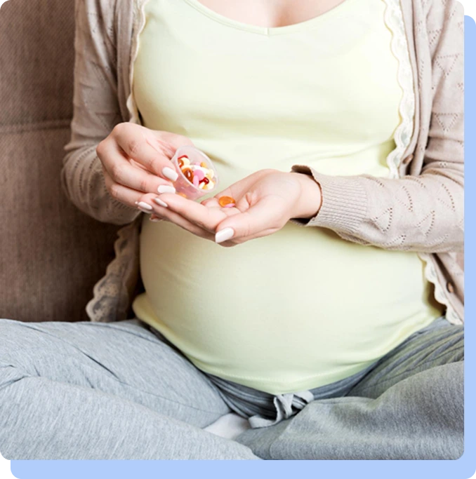 A close-up of a pregnant woman sitting down, holding a small cup of multi-colored supplement capsules in one hand and a single pill in the other.