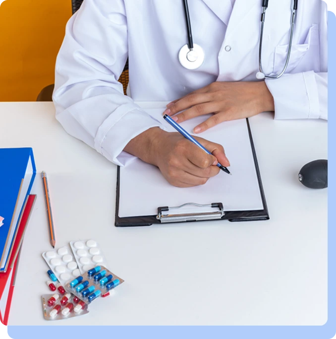 A doctor in a lab coat sitting at a desk, writing on a clipboard next to several blister packs of capsules and a stethoscope.