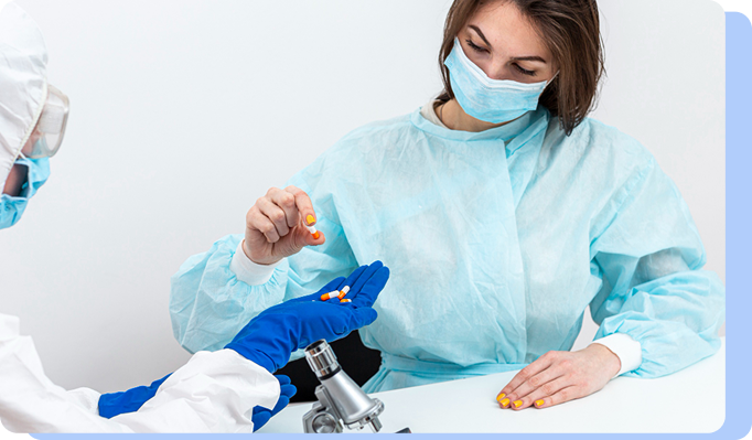 Two healthcare professionals in full surgical gowns, masks, and gloves working together; one is carefully dispensing small orange capsules into the blue-gloved hand of the other.